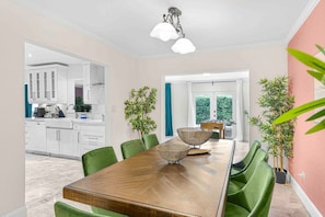 A modern dining area with a wooden table, green chairs, and decorative bowls. There is a kitchen visible in the background with white cabinets and a blue curtain.
