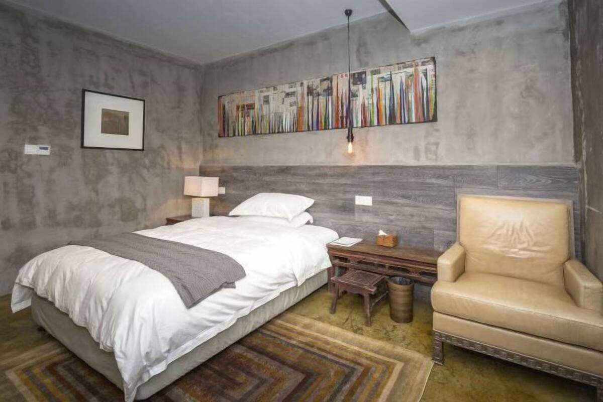 A bedroom with a bed, a chair, a nightstand, a lamp, and a wall art piece.