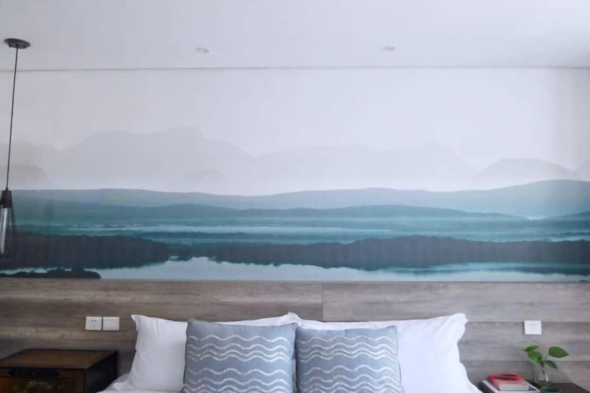 A bedroom with a bed, pillows, a nightstand, a lamp, and a wall with a painting.