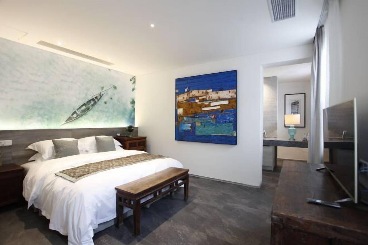 A modern bedroom with a large bed, a nightstand, a bench, a TV, and a painting on the wall.