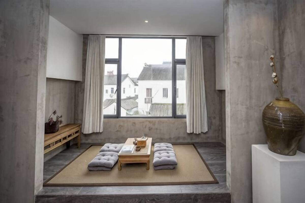 A room with a large window, a tatami mat, a low wooden table, and a vase on a pedestal.