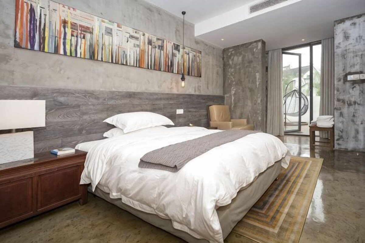 A modern hotel room with a large bed, a nightstand, a chair, and a view of the outdoors.