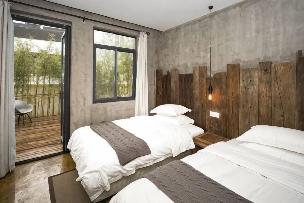 A bedroom with two beds, a wooden headboard, a large window, and a view of greenery outside.