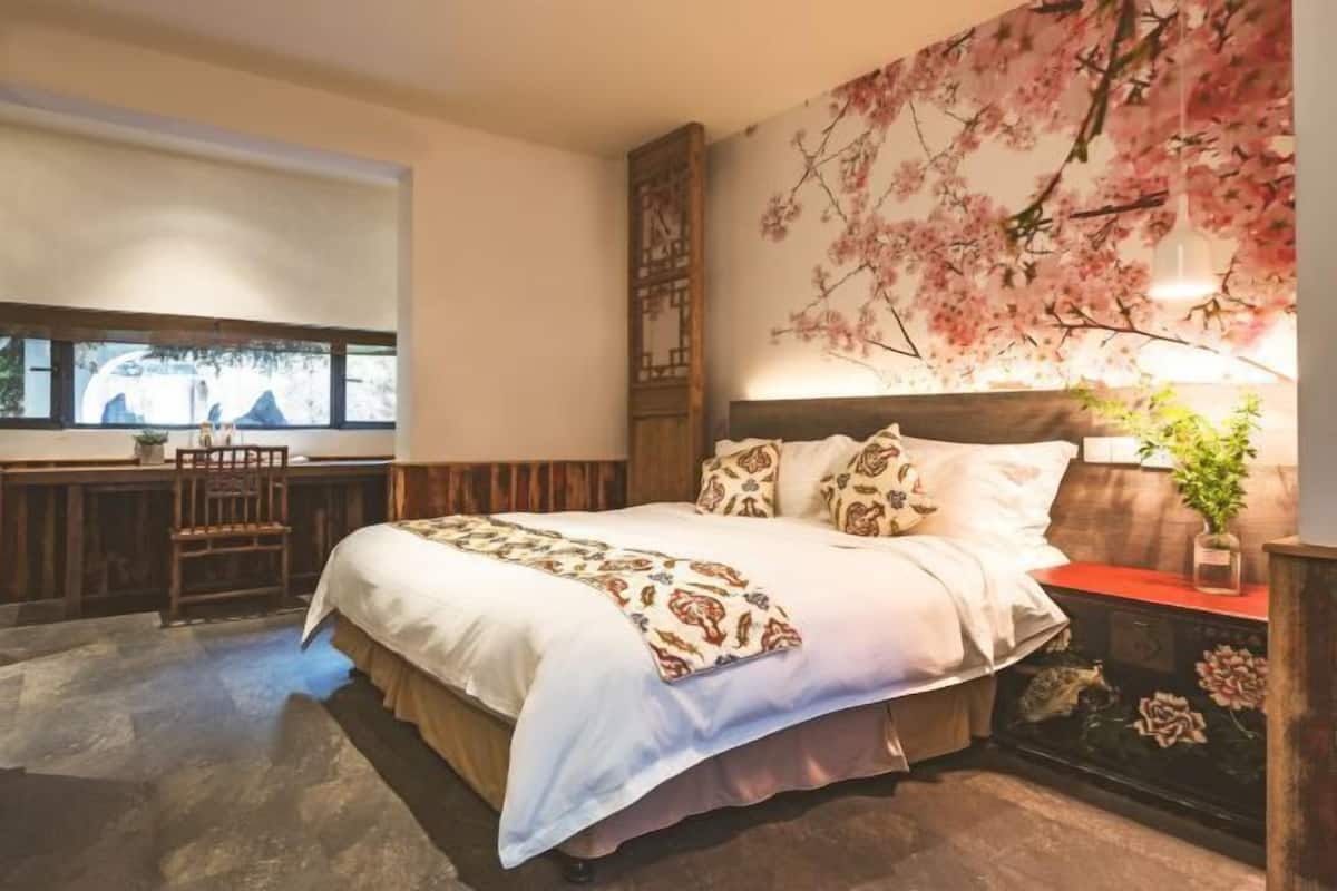 A bedroom with a large bed, a window with a view, a wooden headboard, and a wall with a floral mural.