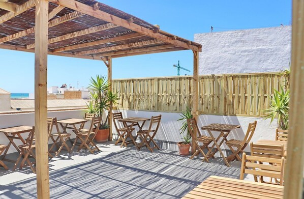 A wooden pergola with tables and chairs, potted plants, and a view of the sea.