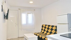 Une petite pièce lumineuse avec une porte blanche, une fenêtre ornée de rideaux blancs, un canapé blanc à motifs noirs et jaunes, une table blanche et un réfrigérateur blanc.