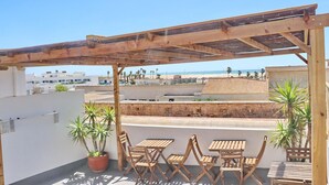 Une terrasse sur le toit, aménagée avec des meubles en bois et des plantes en pot, offrant une vue sur une zone côtière avec des bâtiments et une plage.