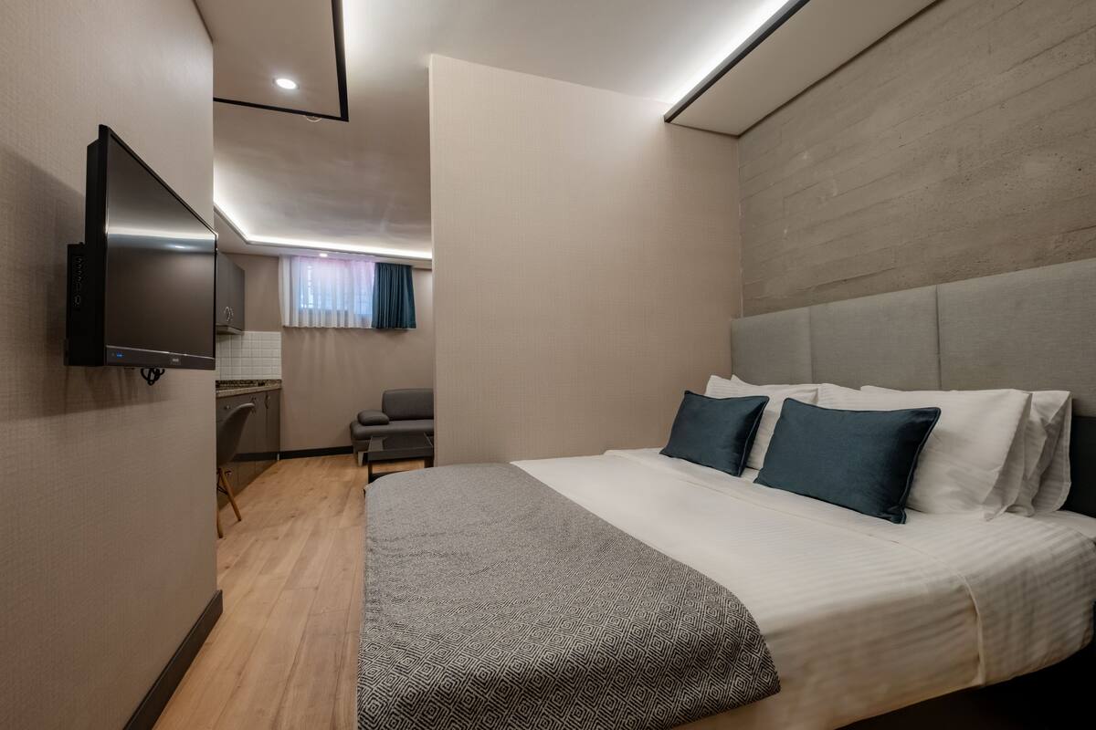A modern hotel room with a large bed, a flat-screen TV, a desk with a chair, and a view of the city through a window.