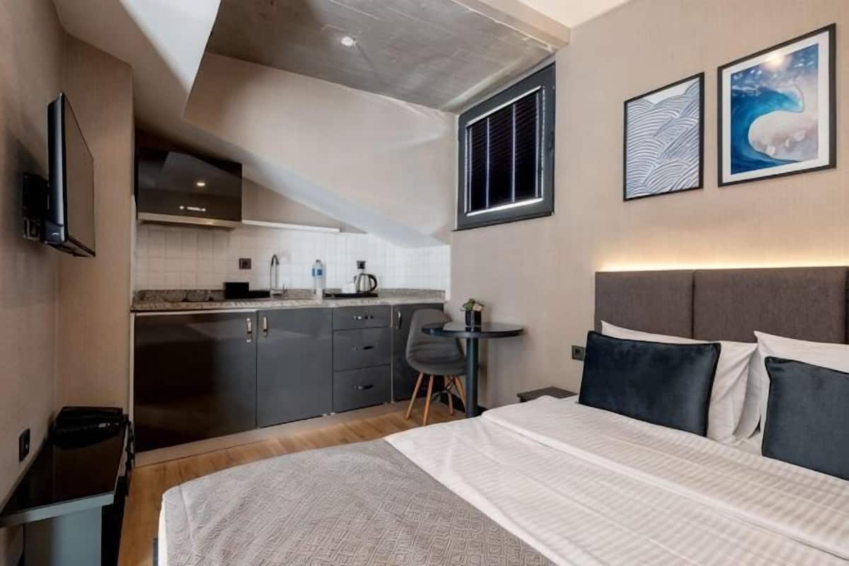 A compact hotel room with a kitchenette, a bed with a headboard, a small table, and two framed pictures on the wall.