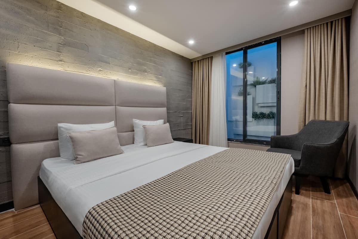 A modern hotel room with a large bed, a headboard, a chair, and a view of the outdoors through a sliding glass door.