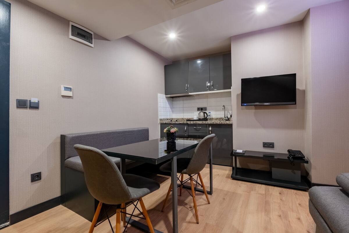 A modern kitchen with a built-in oven, a flat-screen TV mounted above the counter, and a dining area with a glass table and two chairs.