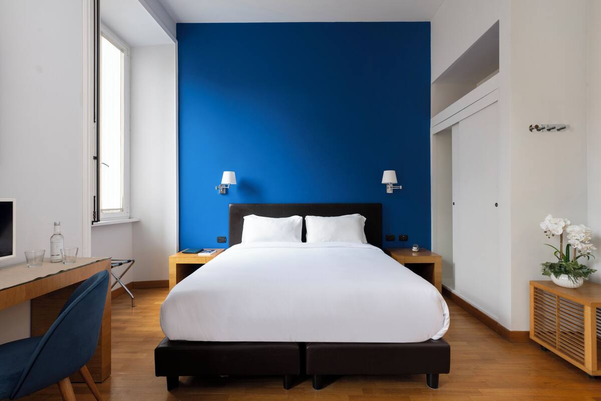 A modern hotel room with a large bed, two bedside tables, a desk with a chair, and a window.