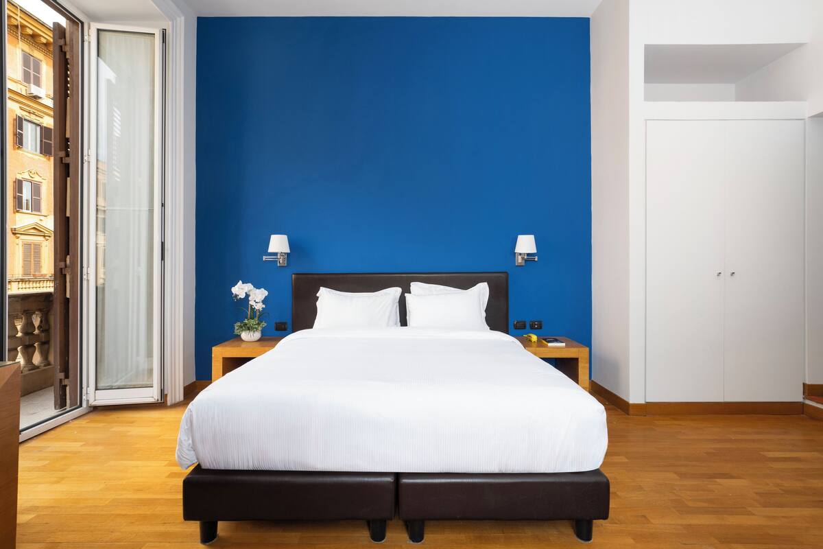 A bedroom with a large bed, two bedside lamps, a nightstand, and a view of a building through a glass door.