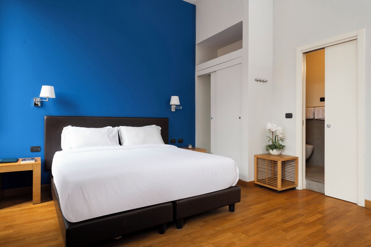 A neatly arranged hotel room with a large bed, bedside tables, a nightstand, a small wooden cabinet, and a bathroom visible through an open door.