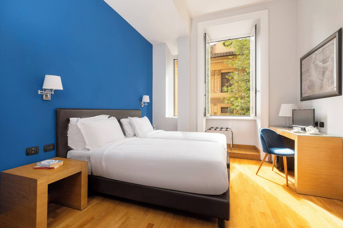 A modern hotel room with a large bed, a desk, and a chair. There are two bedside tables and a window with a view of greenery.