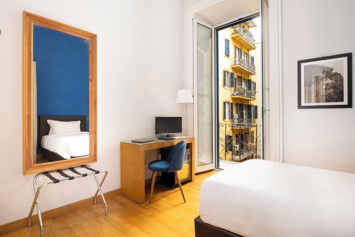A hotel room with a bed, a desk with a computer, a chair, a window with a view of a building, and a framed picture on the wall.