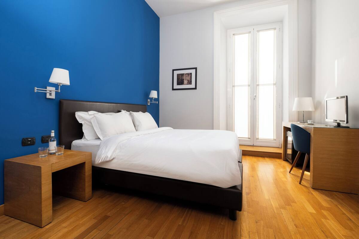 A modern hotel room with a wooden floor, a bed with white linens, a nightstand with a lamp, and a desk with a computer.