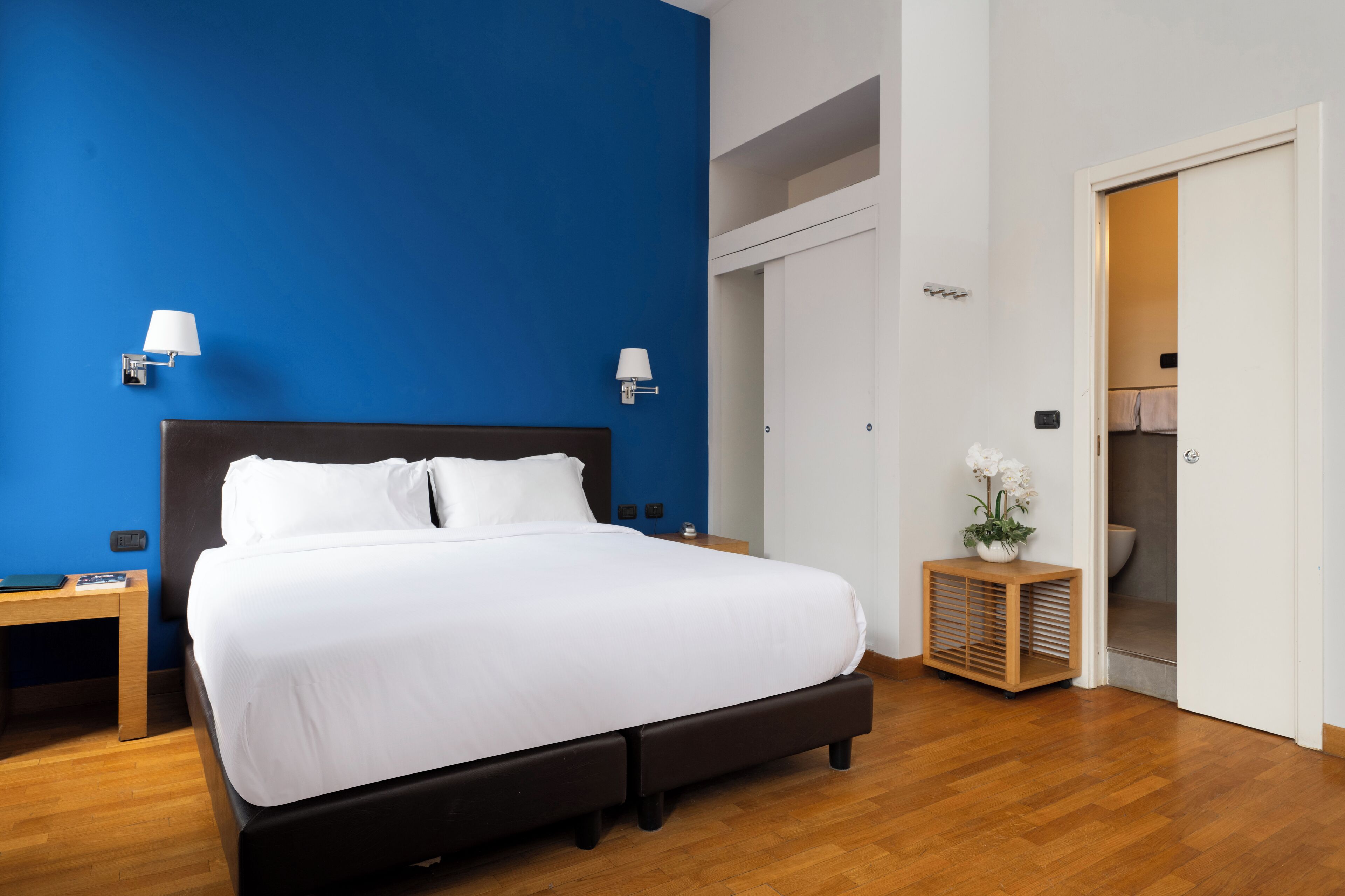 A neatly arranged hotel room with a large bed, bedside tables, a nightstand, a small wooden cabinet, and a bathroom visible through an open door.