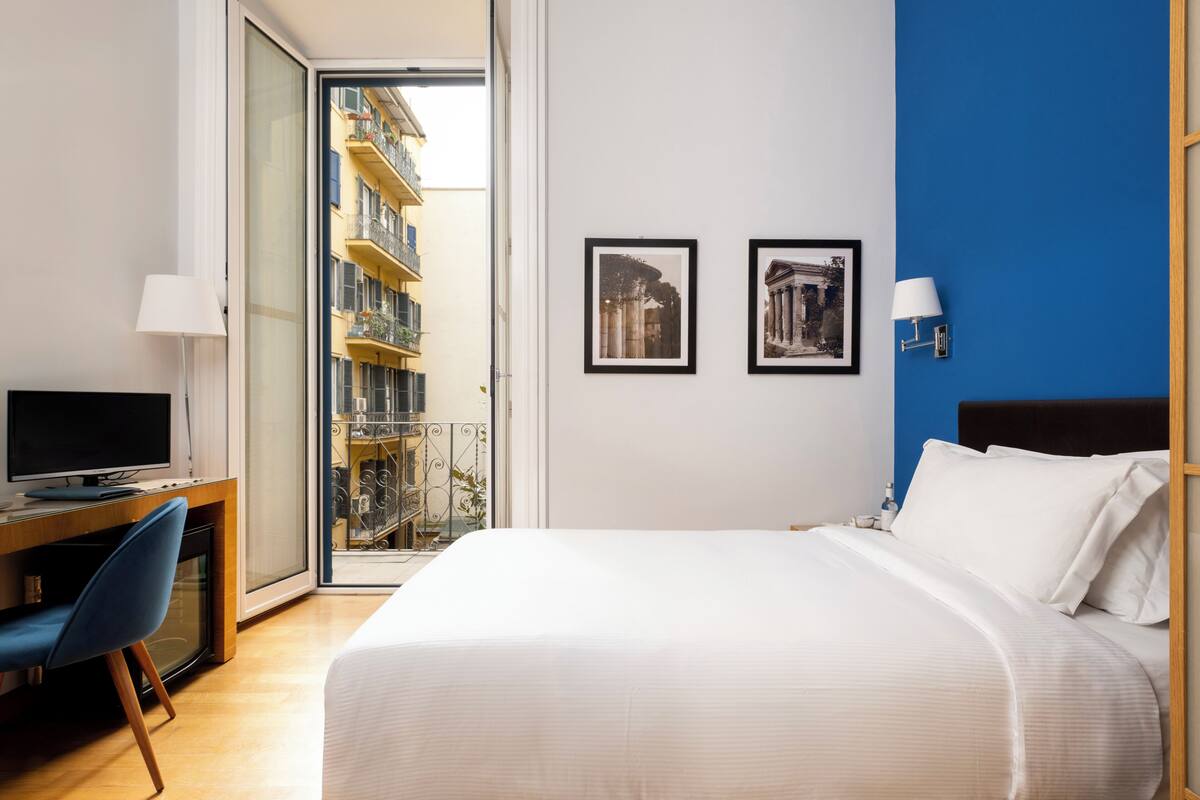 A hotel room with a bed, a desk with a TV, a chair, and two framed pictures on the wall.