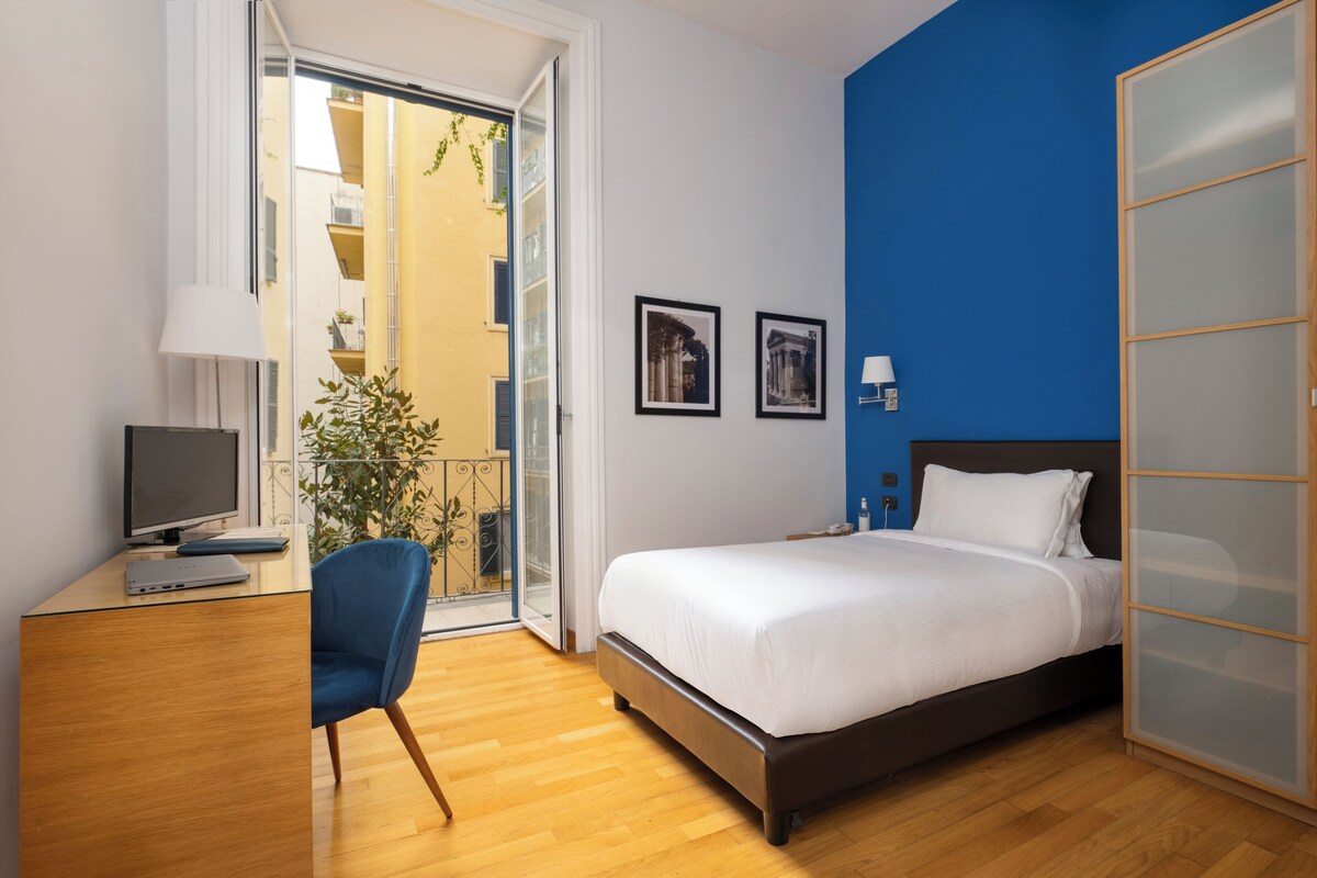 A bedroom with a bed, a desk with a computer, a chair, and a window with a view of buildings.