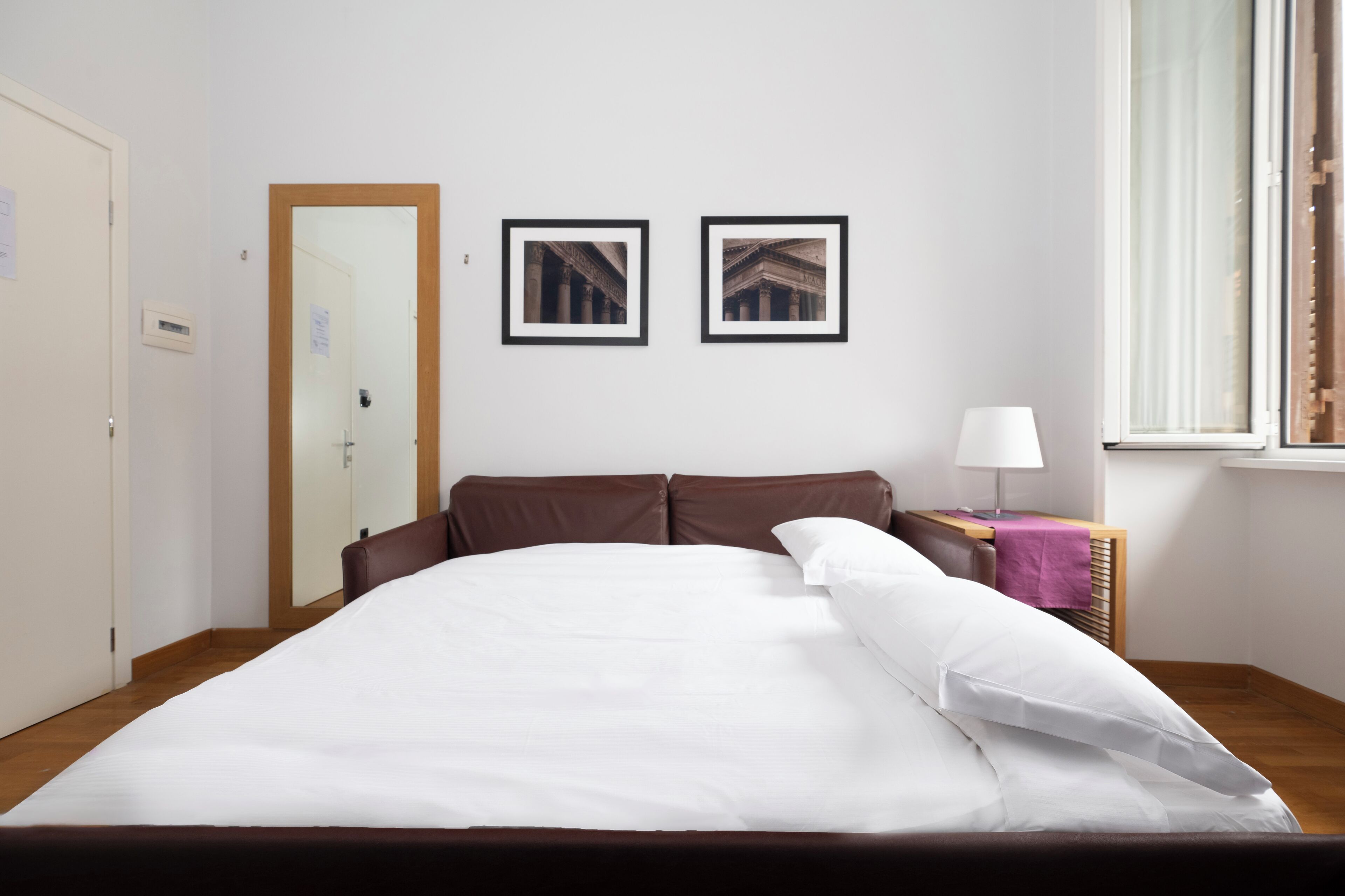 A neatly made bed in a hotel room with a wooden headboard, a bedside table with a lamp, and two framed pictures on the wall.