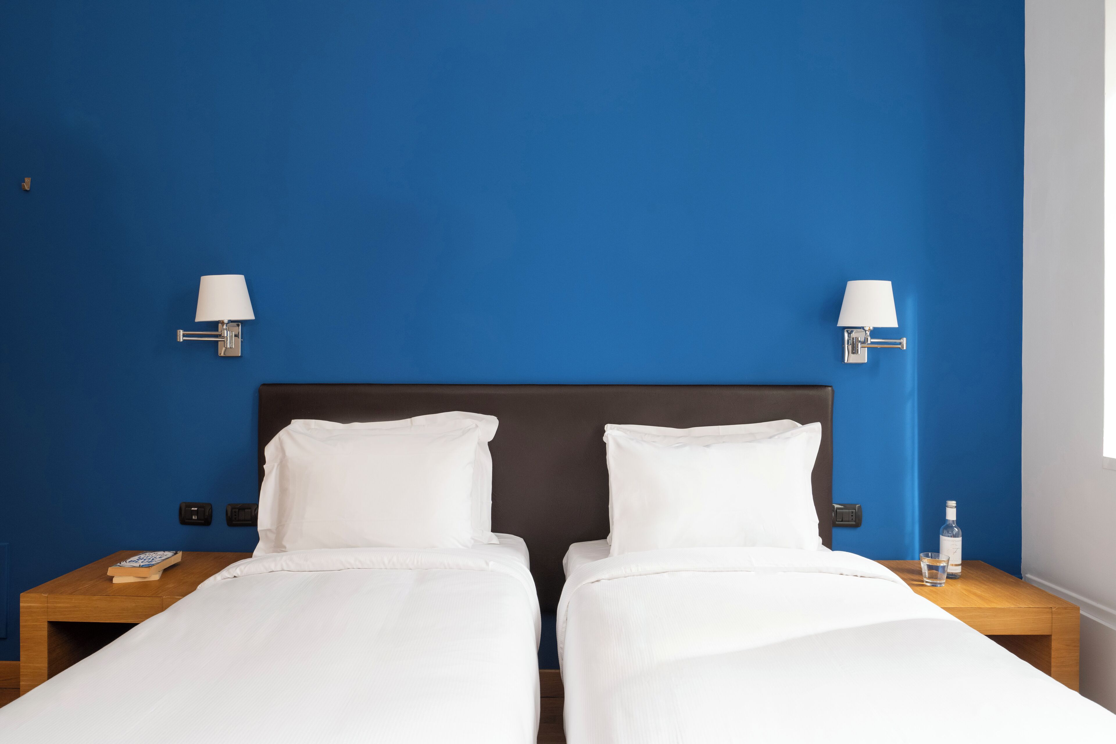 A double bed with white bedding, two bedside tables, a nightstand with a book, and a bottle of water.