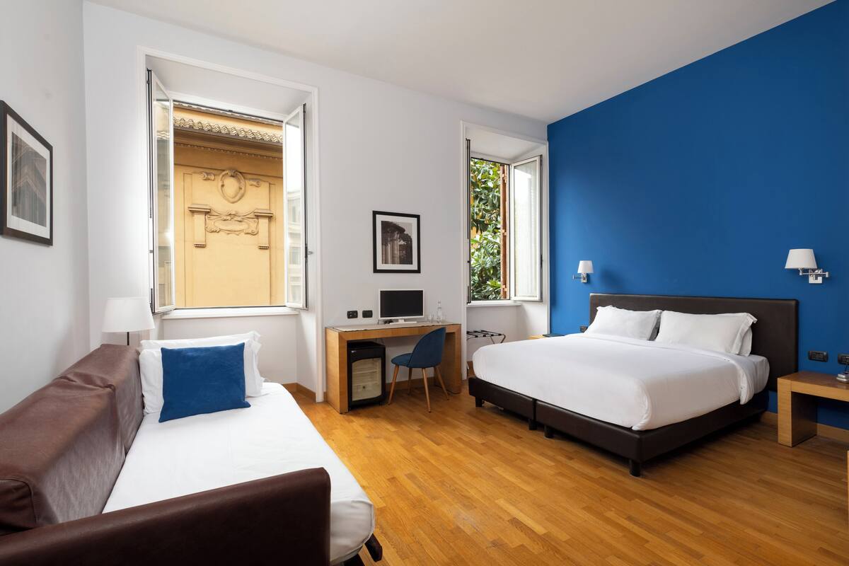 A hotel room with a bed, a desk, a chair, a small refrigerator, and a nightstand.