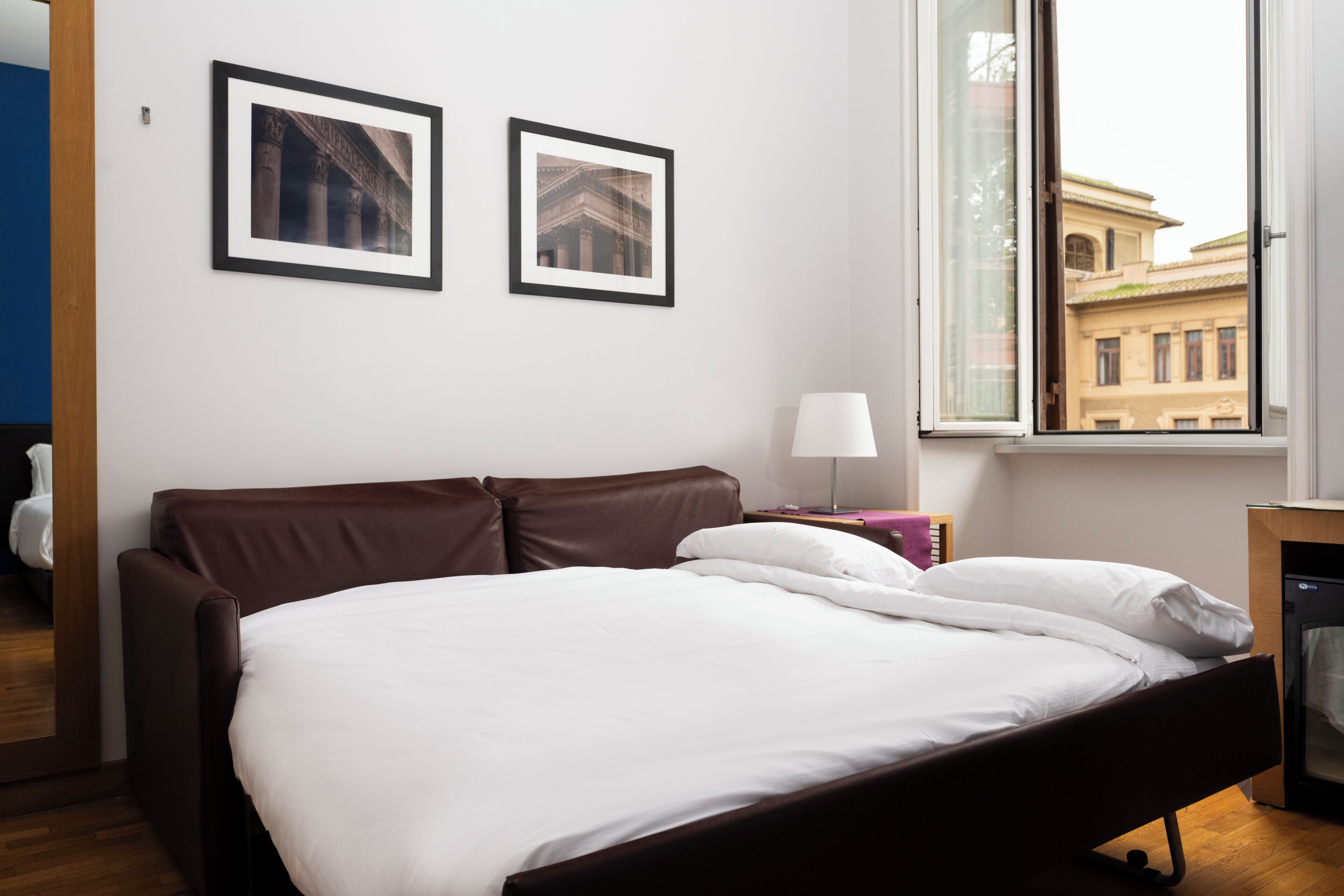 A modern hotel room with a large bed, two framed pictures on the wall, a window with a view of buildings, and a bedside table with a lamp.