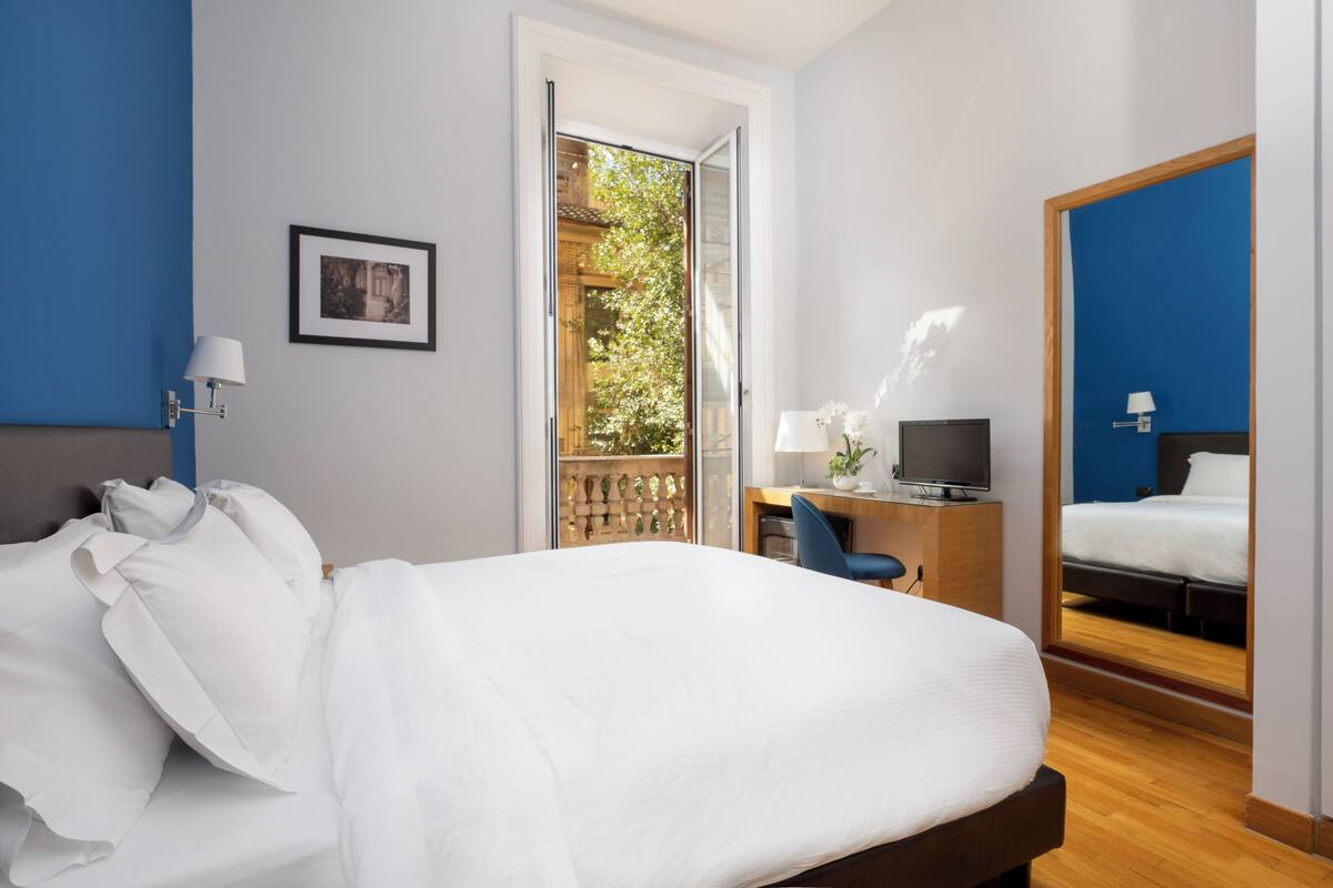 A hotel room with a bed, desk, TV, and a balcony with a view of greenery.