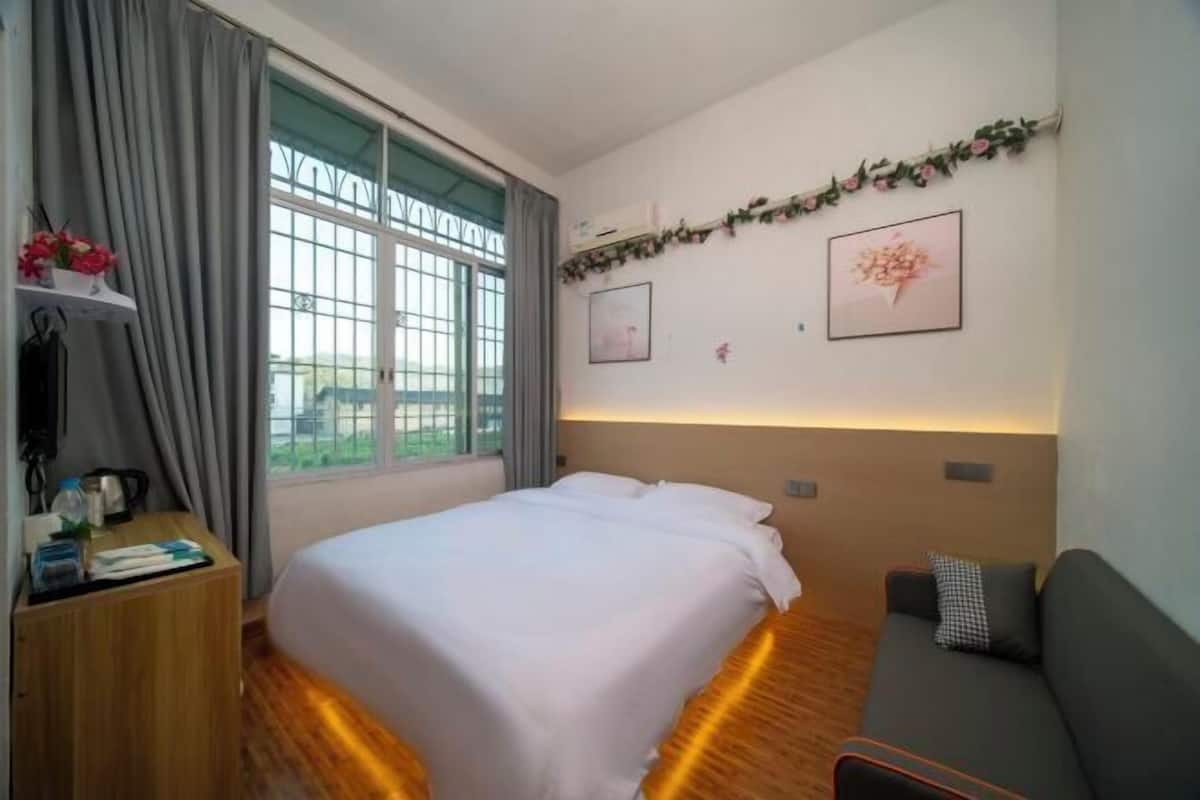 A hotel room with a bed, a sofa, a wooden nightstand, a window with curtains, and wall-mounted artwork.