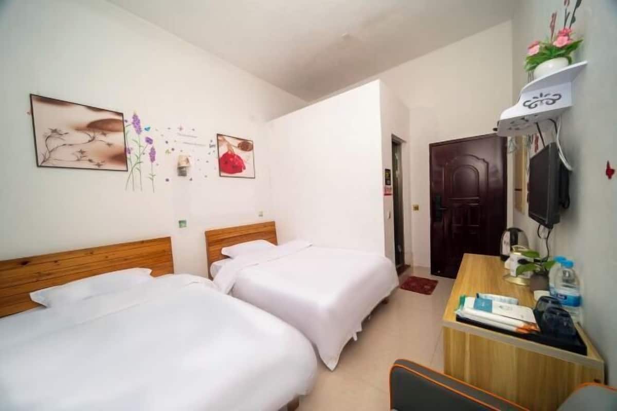 A hotel room with two single beds, a wooden headboard, a desk with a chair, a TV mounted on the wall, and a door leading to another room.