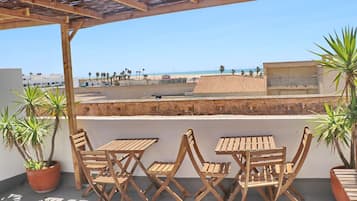 Une terrasse dotée de tables et de chaises en bois, de plantes en pot, et offrant une vue sur la plage et le paysage urbain.