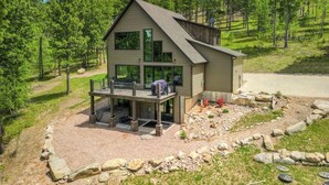 Ein modernes, zweistöckiges Haus mit einer großen Terrasse, umgeben von einer naturnahen Landschaft und einer Schotterauffahrt.