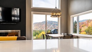 A modern dining area with a large table, chairs, a fireplace, and a view of autumn foliage through large windows.
