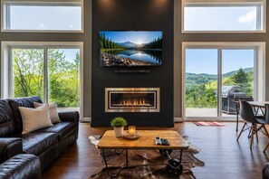 A modern living room with a fireplace, a leather sofa, a coffee table, and a large TV screen displaying a scenic landscape. - Incredible view & Spa | L'Odace (Saint-Côme)