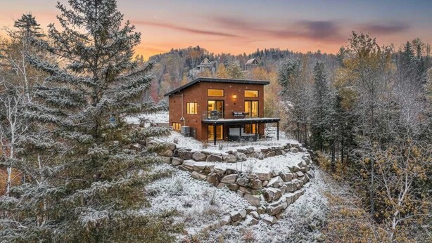 A modern cabin with a stone foundation, a patio, and a view of the surrounding forest.