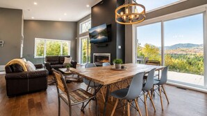 A modern living room with a fireplace, a dining area with a wooden table, and large windows offering a scenic view.
