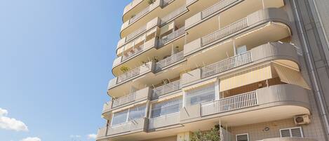 A multi-story residential building with balconies, tiled exterior, and a clear sky.