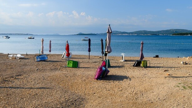 A pebbled beach with beach chairs, umbrellas, and a row of luggage. Boats are visible in the water.