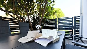 A book and a hat on a wooden table outdoors with a metal fence and trees in the background.