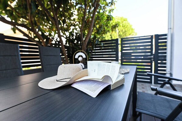 A book and a hat on a wooden table outdoors with a metal fence and trees in the background.