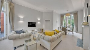 A modern living room with a sofa, a coffee table, and a TV. There is a dining area with a table and chairs. - Cosy One Bedroom Behind Harrods (Greater London)