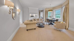 Een ruime woonkamer met een bank, fauteuils en een salontafel. Er is een eethoek met een tafel en stoelen, en een groot raam met gordijnen.