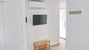 Et værelse med en hvid dør, en fastmonteret aircondition, et fladskærms-tv, en træbænk og en lyskontakt på væggen.