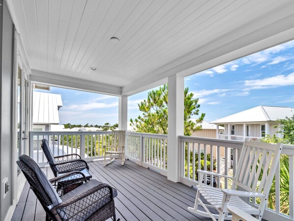 A covered porch with two wicker chairs and a rocking chair, overlooking a residential area with houses and trees.