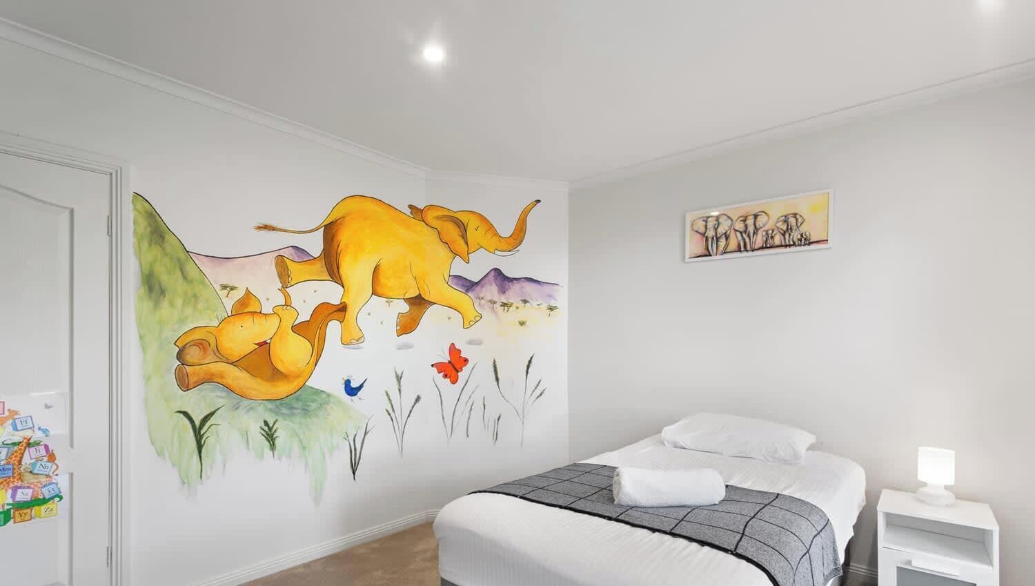 A bedroom with a large wall mural, a bed with white linens, a bedside table with a lamp, and a small artwork above the bed.