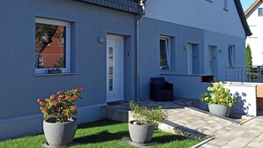 Una casa de dos pisos con un balcón, una puerta y una ventana. Hay plantas en macetas y una silla afuera.