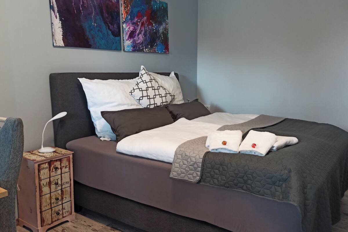 Een netjes opgemaakt bed met een grijze dekbedovertrek, meerdere kussens en een decoratieve doos met tekst aan de zijkant.