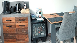 Una kitchenette compacta con cafetera, hervidor y un estante de almacenamiento. También se observa un refrigerador pequeño y una mesa con una silla.