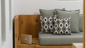 Une pièce moderne avec un banc en bois, un canapé gris, des coussins à motifs, un ordinateur portable et une lampe noire fixée au mur.